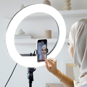 A woman in a hijab uses a smartphone with a ring light for recording indoors.
