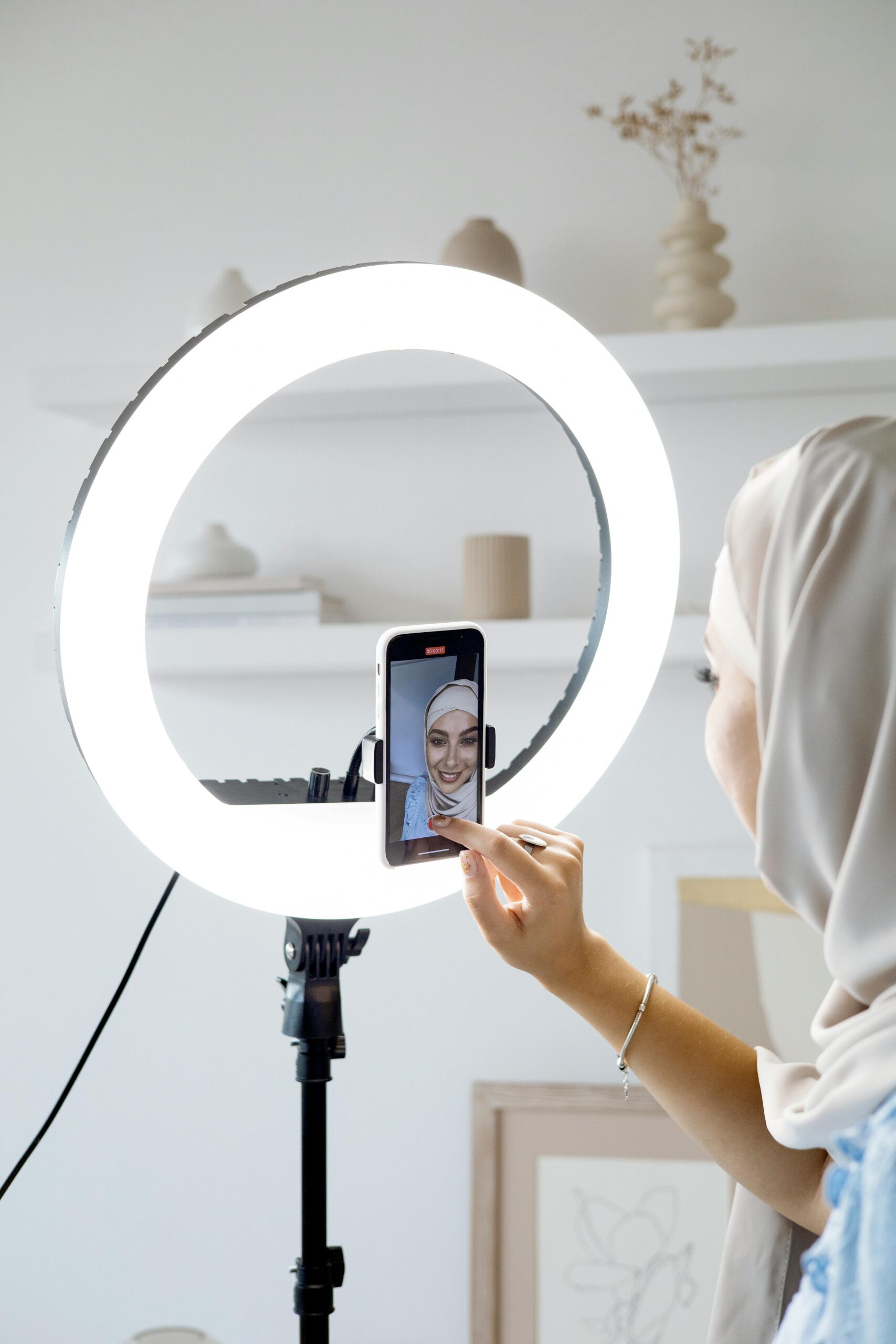 A woman in a hijab uses a smartphone with a ring light for recording indoors.